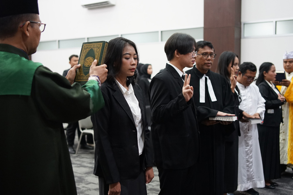Group of formally dressed participants standing in a ceremony; a man in green robes holds a large book as others raise a peace sign or hold items.