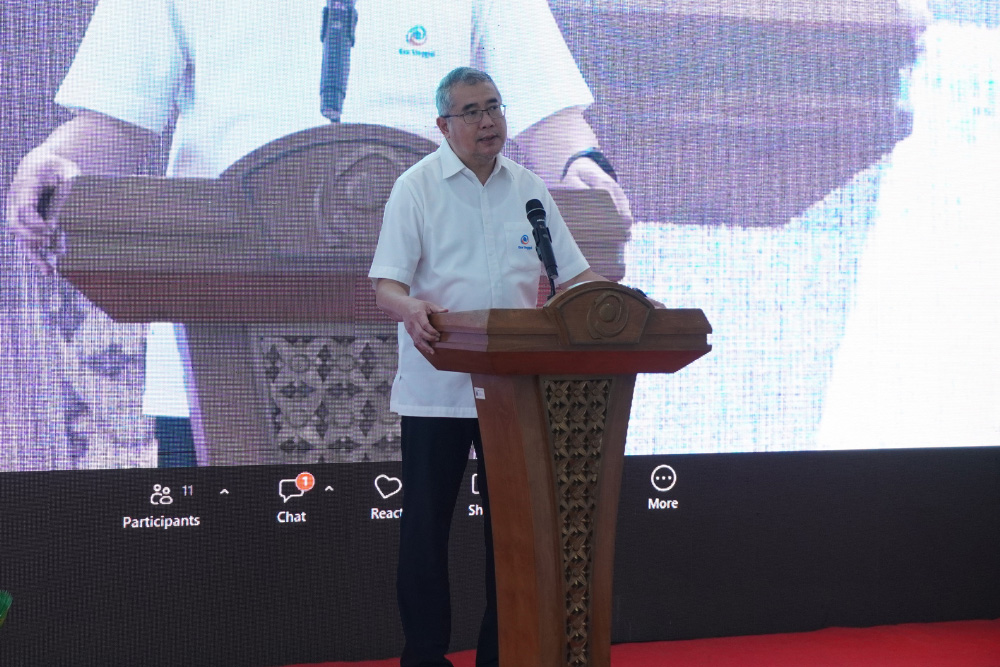 Man in a white shirt speaks at a carved wooden podium on a stage with a large screen behind him with an image projection.