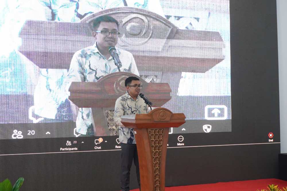 Man in a floral shirt delivers a speech from a wooden podium on a stage, with a large screen showing his image behind him.