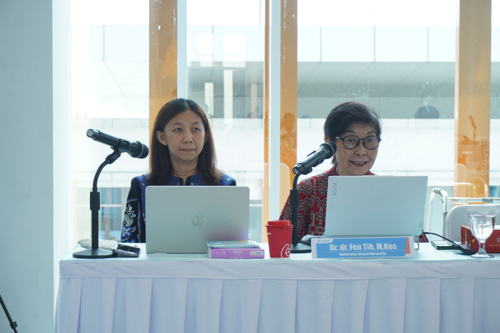 Two women sit at a conference table with laptops and microphones, presenting at a panel.