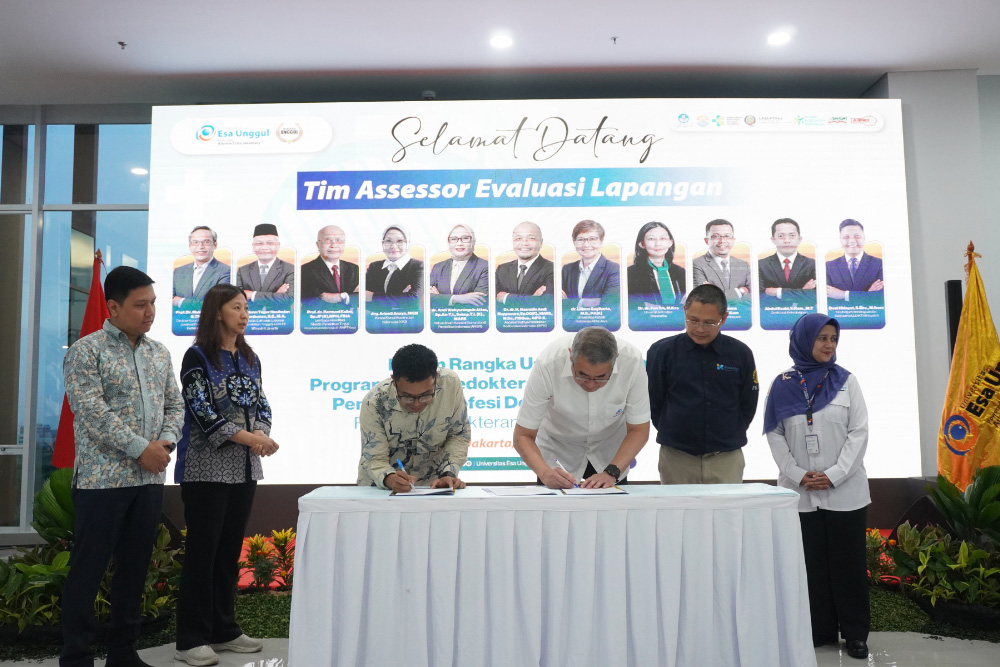 Six people stand around a table as two men sign documents during a formal signing ceremony, with a large banner behind them.