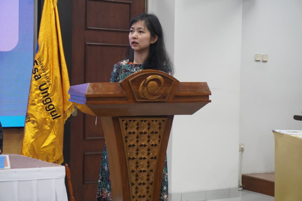 Woman giving a speech at a wooden podium in a conference room, with a yellow flag in the background.