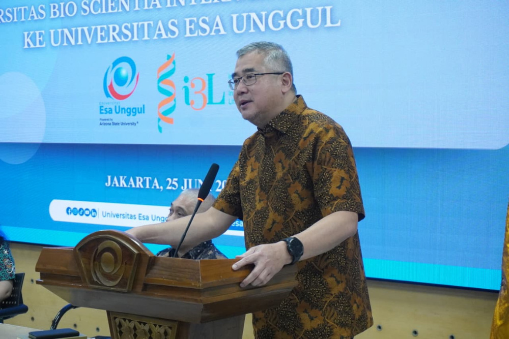 Man in brown batik shirt speaks at a wooden podium with a microphone, in front of a blue event banner showing Jakarta information and logos.
