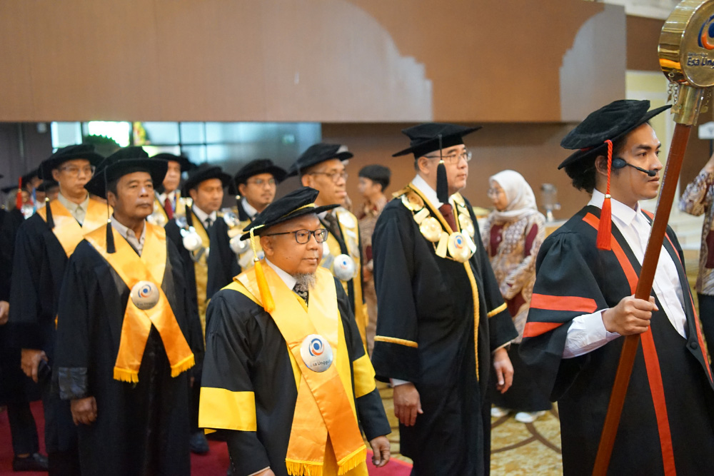 Group of graduates in black gowns and yellow stoles marching in a ceremony indoors.