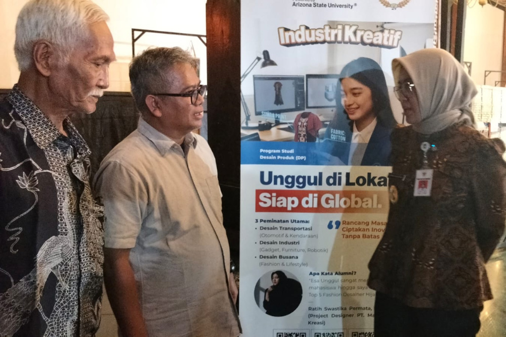 Three adults stand together and discuss a promotional poster for Industri Kreatif at a workshop or expo.