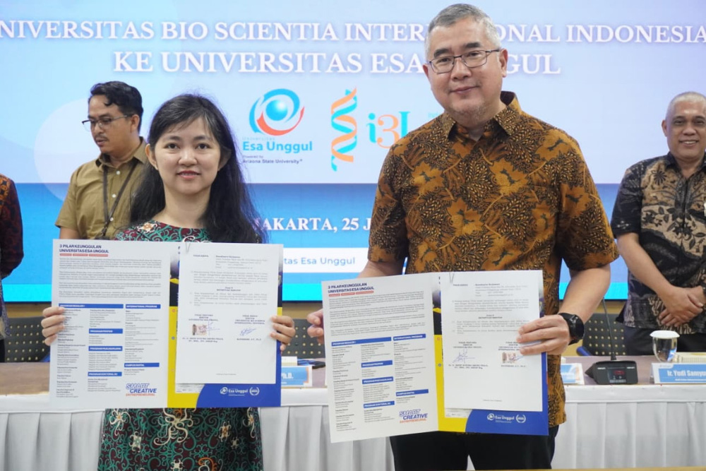 Man and woman hold displayed documents during a formal signing ceremony, with a blue banner backdrop and other attendees nearby in batik shirts.