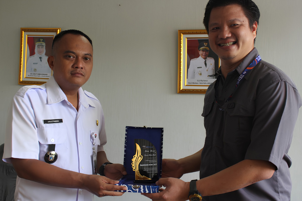 Two men stand indoors as one in a white uniform presents a glass trophy to a man in a dark shirt, framed portraits on the wall behind them.