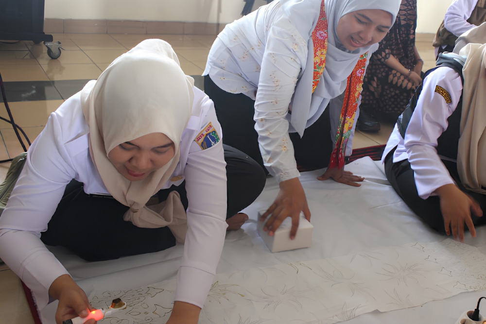 Group of students in white uniforms and hijabs kneeling on a mat, drawing on a long sheet of paper spread on the floor in a classroom.